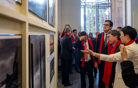 Várias pessoas em espaço interior apreciam uma exposição de fotografias. Todas as pessoas usam cachecol vermelho.