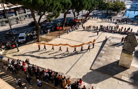 Fotografia, em plano superior e elevado, de praça. Na praça estão algumas dezenas de pessoas que assistem a uma atuação da dança do dragão. Do lado direito vê-se uma estátua. À esquerda e ao fundo identificam-se viaturas e árvores.