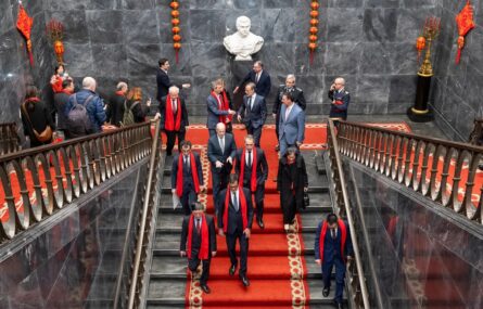 Perto de duas dezenas de pessoas descem escadaria (com tapete vermelho). Nas paredes estão pendurados motivos alusivos ao Ano Novo Chinês. Quase todas as pessoas usam cachecol vermelho.
