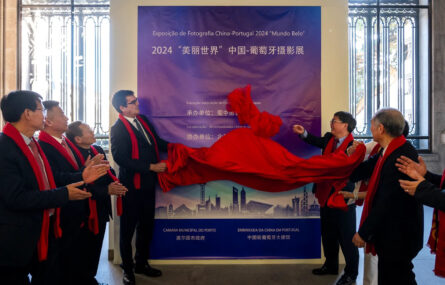 Duas pessoas retiram pano vermelho que cobria enorme cartaz (em tons de azul) alusivo à exposição de fotografia China-Portugal. Ao lado outras pessoas aplaudem. Todas usam cachecol vermelho.