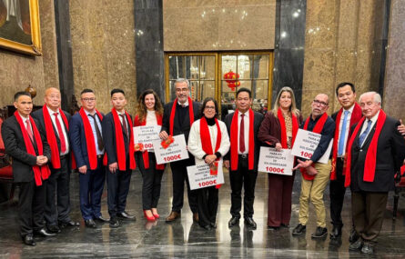 Fotografia de 12 pessoas, alinhadas, em espaço interior. Cinco das pessoas seguram cartazes que indicam “donativo para as associações de solidariedade”. Todas usam cachecol vermelho.