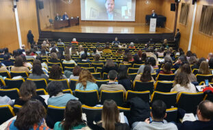 Fotografia de auditório. Perto de duas centenas de pessoas sentadas na audiência. Ao fundo um palco com mesa de abertura do congresso e púlpito. Duas pessoas sentadas junto da mesa. Uma pessoa, em pé, a falar junto do púlpito. Ao centro, em projeção em tela, um outro interveniente na cerimónia de abertura (em transmissão digital).