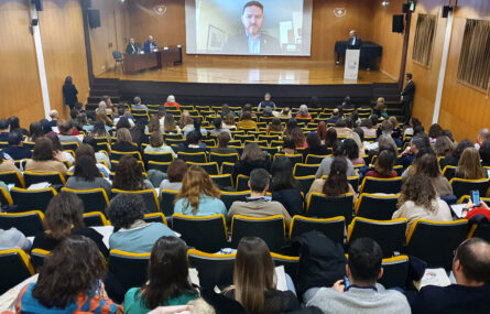 Fotografia de auditório. Perto de duas centenas de pessoas sentadas na audiência. Ao fundo um palco com mesa de abertura do congresso e púlpito. Duas pessoas sentadas junto da mesa. Uma pessoa, em pé, a falar junto do púlpito. Ao centro, em projeção em tela, um outro interveniente na cerimónia de abertura (em transmissão digital).