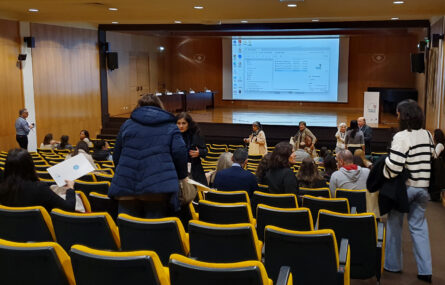 Aspeto geral de auditório (ainda com poucas pessoas). Ao fundo um palco com uma mesa e uma projeção em tela. Algumas pessoas, em pé, estão a conversar.