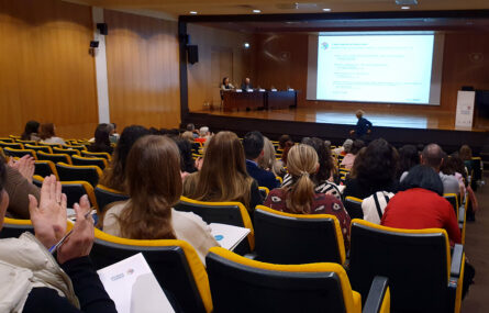 Aspeto geral de auditório (já parcialmente ocupado com pessoas sentadas). Ao fundo um palco com uma mesa e uma projeção em tela. No palco estão duas pessoas sentadas junto de uma mesa.