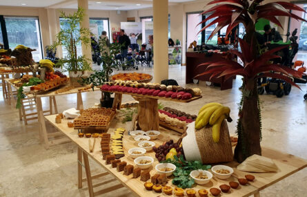 Fotografia de espaço interior com várias mesas em madeira. Todas as mesas têm pousados vários produtos comestíveis (e algumas plantas como decoração). Ao fundo algumas pessoas.