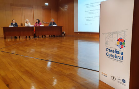 Palco de auditório com uma mesa do lado esquerdo. Junto da mesa estão quatro pessoas sentadas. Do lado direito da imagem um púlpito com um cartaz do congresso de paralisia cerebral. Vê-se ainda, parcialmente, projeção em tela ao centro da imagem.