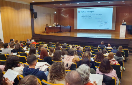 Aspeto geral de auditório (já consideravelmente ocupado com pessoas sentadas). Ao fundo um palco com uma mesa e uma projeção em tela. No palco, junto da mesa, estão duas pessoas sentadas. Outra pessoa está a falar, junto de um púlpito (do lado direito da imagem).