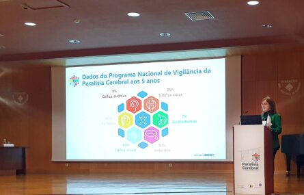 Pessoa a falar junto de um púlpito. Mais ao fundo está uma tela gigante com uma projeção de dados do Programa Nacional de Vigilância da Paralisia Cerebral aos 5 anos.