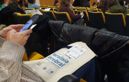 Uma pessoa, sentada, pesquisa informações no telemóvel. Pousado nas pernas tem um saco com material referente ao congresso. Ao fundo mais pessoas igualmente sentadas.