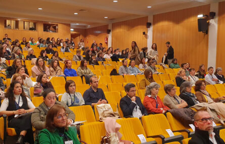 Fotografia de auditório. Várias dezenas de pessoas sentadas em cadeiras (todas em tom amarelo escuro). Algumas pessoas de pé a assistir.