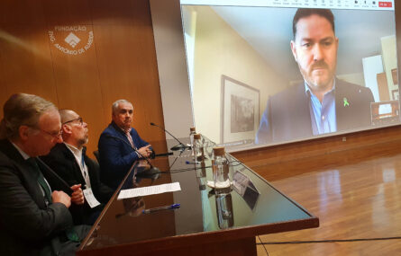 Do lado esquerdo da imagem estão três pessoas sentadas junto de uma mesa. À direita, em projeção em ecrã, está uma outra pessoa.