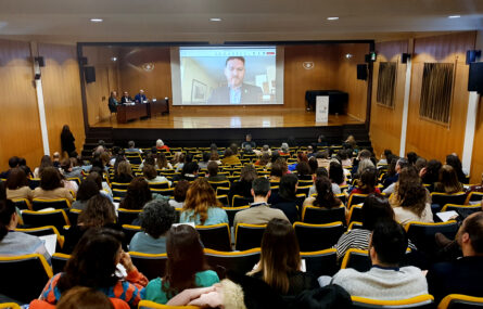 Aspeto geral de auditório (já consideravelmente ocupado com pessoas sentadas). Ao fundo um palco com uma mesa e uma projeção em tela. No palco, junto da mesa, estão três pessoas sentadas. Na projeção em tela surge ainda outra pessoa.