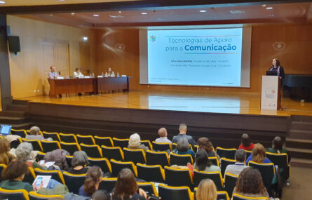 Aspeto geral do auditório com várias pessoas sentadas nas primeiras filas. Palco com mesa e três pessoas. Púlpito com uma pessoa a falar. Projeção em ecrã gigante, ao centro.