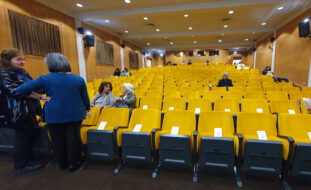 Fotografia de auditório praticamente vazio. Algumas poucas pessoas, sentadas ou em pé, conversam.