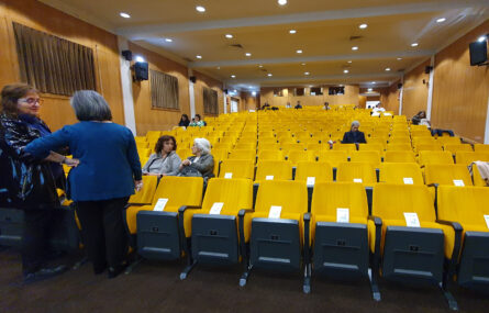 Fotografia de auditório praticamente vazio. Algumas poucas pessoas, sentadas ou em pé, conversam.
