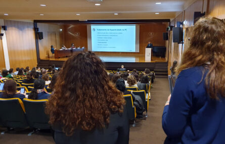 Em primeiro plano duas pessoas, de costas. Mais ao fundo da imagem aspeto geral do auditório com várias dezenas de pessoas sentadas. Palco com mesa e três pessoas. Púlpito com uma pessoa a falar. Projeção em ecrã gigante, ao centro.