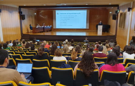 Aspeto geral do auditório com várias dezenas de pessoas sentadas. Em primeiro plano está uma pessoa, sentada, a utilizar um computador portátil. Palco com mesa e três pessoas. Púlpito com uma pessoa a falar. Projeção em ecrã gigante, ao centro.