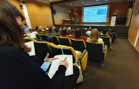 Aspeto geral do auditório com várias dezenas de pessoas sentadas. Em primeiro plano está uma pessoa, sentada, a escrever numa folha de papel e a consultar algo no telemóvel. Palco com mesa e duas pessoas. Púlpito com uma pessoa a falar. Projeção em ecrã gigante, ao centro.