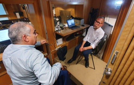 Imagem de pequena sala de apoio técnico em auditório. Duas pessoas sentadas a conversar. Diverso equipamento técnico espalhado.