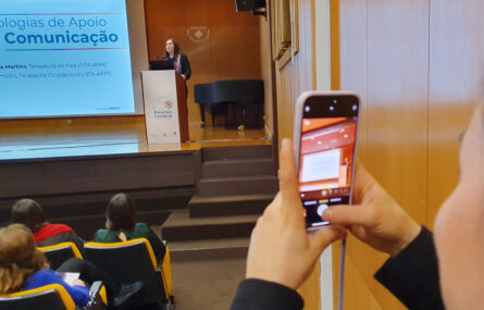 Em primeiro plano uma pessoa utiliza o telemóvel para fazer uma fotografia. Ao fundo da imagem parte do palco e uma pessoa a falar junto de um púlpito. Vê-se, ainda que parcialmente, projeção em ecrã gigante.