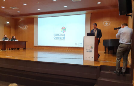 Palco do auditório. Do lado esquerdo mesa com duas pessoas. Ao centro projeção em ecrã gigante. À direita pessoa a falar junto de púlpito e uma outra pessoa, encostada à parede, a fazer uma fotografia (com máquina fotográfica).