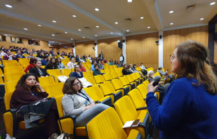 Pessoa com microfone na mão a falar para outras dezenas de pessoas sentadas no auditório.