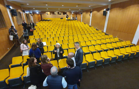 Fotografia, ampla, de auditório praticamente vazio. Algumas pessoas conversam.