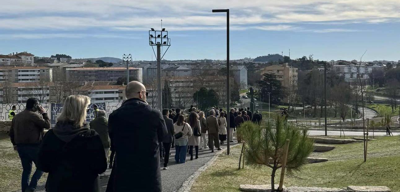 Grupo de pessoas a caminhar em percurso num amplo espaço verde. Edifícios ao fundo da imagem.