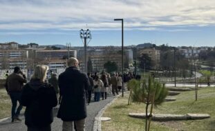 Grupo de pessoas a caminhar em percurso num amplo espaço verde. Edifícios ao fundo da imagem.