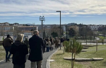 Grupo de pessoas a caminhar em percurso num amplo espaço verde. Edifícios ao fundo da imagem.
