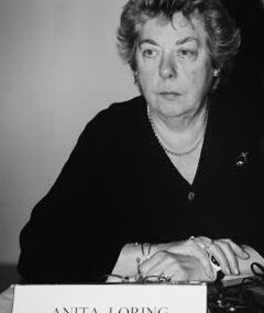Fotografia, em tons cinza, de pessoa junto de uma mesa. Tem as mãos cruzadas e apoiadas em cima da mesa. À sua frente aparece uma placa identificativa com o nome Anita Loring.
