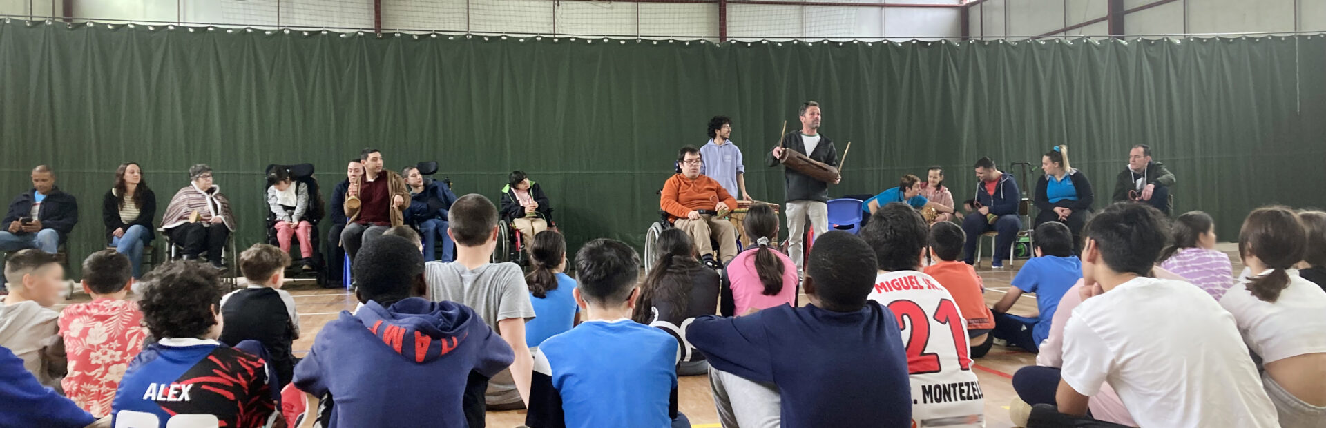 Fotografia em ginásio. Perto de duas dezenas de jovens alunos, de costas, sentados no chão. Mais ao fundo, junto de uma cortina verde, mais de uma dezena representantes da APPC. Os rostos visíveis de crianças foram desfocados.
