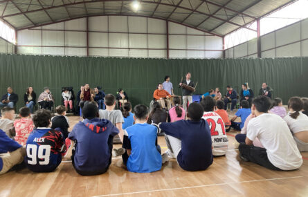 Fotografia em ginásio. Perto de duas dezenas de jovens alunos, de costas, sentados no chão. Mais ao fundo, junto de uma cortina verde, mais de uma dezena representantes da APPC. Os rostos visíveis de crianças foram desfocados.