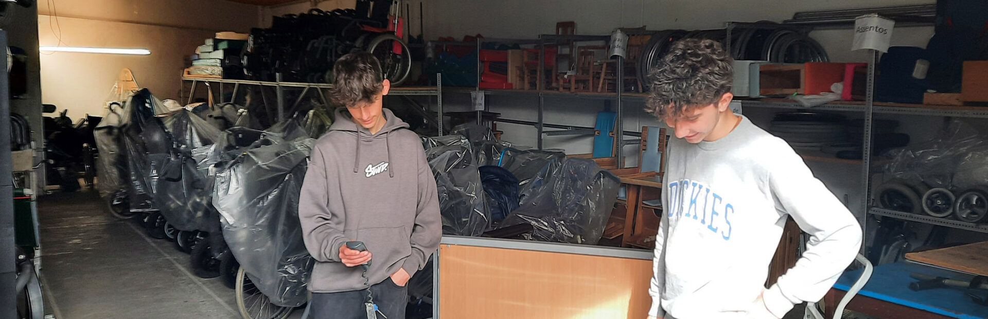 Em espaço interior, que aparenta ser um armazém, dois jovens testam o comando de uma cama articulada. Mais ao fundo da imagem, no chão e em prateleiras, estão várias dezenas de produtos de apoio devidamente armazenados.