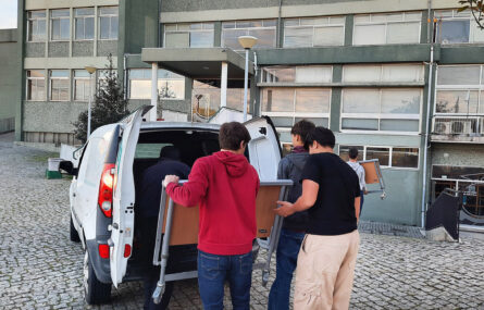 Três jovens junto de uma viatura que se encontra com as portas traseiras abertas. Estão a colocar um equipamento no interior da viatura. Mais ao fundo da imagem outro jovem segura o que aparenta ser parte complementar do referido equipamento.