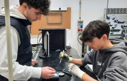 Dois jovens junto de uma mesa. Um deles usa ferro de soldar. A mesa tem ainda um computador e um ecrã pousados.