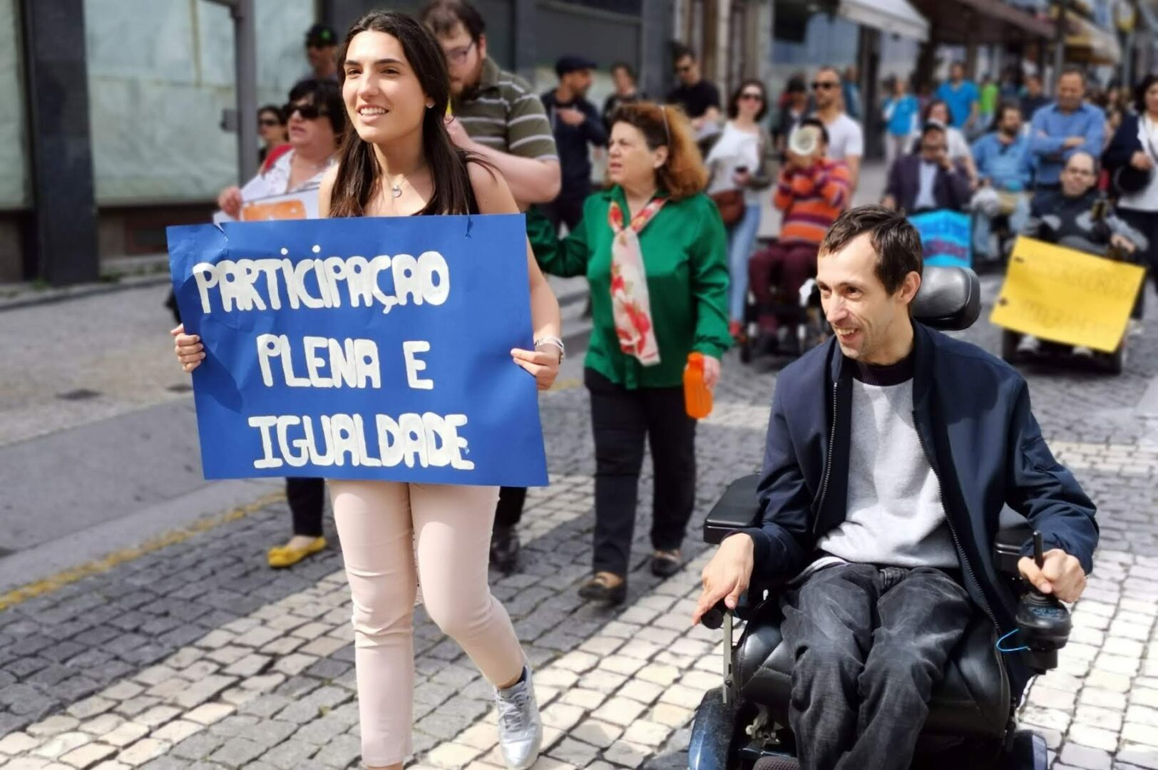 Várias pessoas transitam em rua de paralelos. À frente do grupo uma pessoa segura um cartaz onde se lê: Participação plena e igualdade.