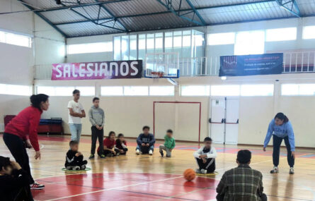 Grupo de crianças, jovens e adultos em ginásio. Desenvolvem uma atividade com uma bola (que aparenta ser de basquetebol). Ao fundo uma lona com a designação Salesianos. Os rostos das crianças e jovens foram digitalmente desfocados.