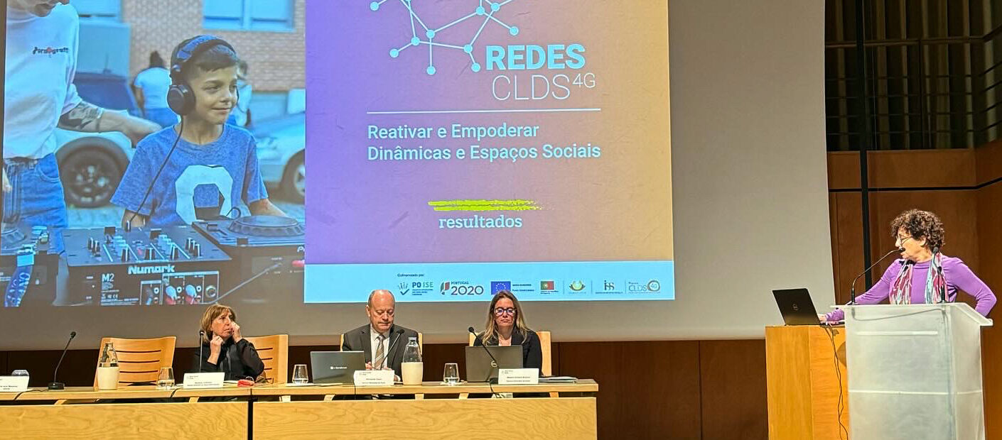 Três pessoas sentadas junto de duas mesas, em cima do palco. Ao fundo, em tela, está projetada uma apresentação. À direita, junto de um púlpito, está uma pessoa a falar.