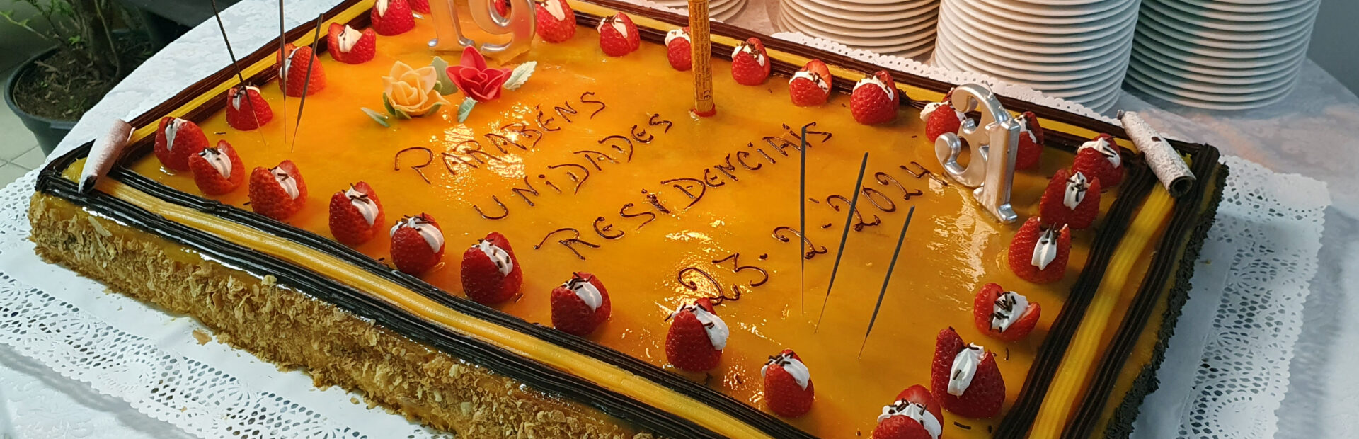 Bolo de aniversário com velas acesas. Em cima da mesa estão, também, várias dezenas de pratos empilhados. No bolo lê-se “Parabéns Unidades Residenciais”.