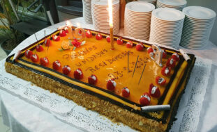 Bolo de aniversário com velas acesas. Em cima da mesa estão, também, várias dezenas de pratos empilhados. No bolo lê-se “Parabéns Unidades Residenciais”.