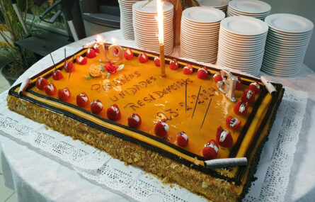 Bolo de aniversário com velas acesas. Em cima da mesa estão, também, várias dezenas de pratos empilhados. No bolo lê-se “Parabéns Unidades Residenciais”.
