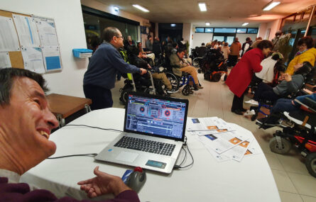 Mais de duas dezenas de pessoas em zona de entrada de edifício. Em primeiro plano uma pessoa utiliza o computador portátil para colocar música ambiente. Ao lado do computador portátil estão pousados alguns panfletos.