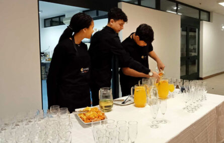 Três jovens junto de uma mesa com inúmeros copos, várias jarras e uma travessa repleta de fatias de laranja já cortadas.