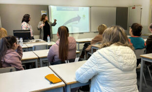 Espaço interior que aparenta ser uma sala de aulas. Várias pessoas sentadas junto de inúmeras mesas. Ao fundo, em pé, outras duas pessoas fazem uma explicação oral com recurso a uma projeção multimédia.