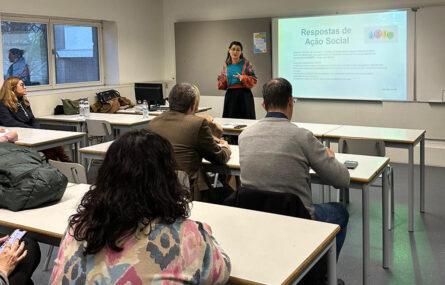 Espaço interior que aparenta ser uma sala de aulas. Várias pessoas sentadas junto de inúmeras mesas. Ao fundo, em pé, uma outra pessoa faz uma explicação oral com recurso a uma projeção multimédia.
