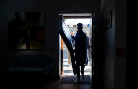 Fotografia, em contraluz, de pessoa a entrar por uma porta aberta. A referida pessoa leva algo nas mãos. Atrás surgem ainda mais pessoas.