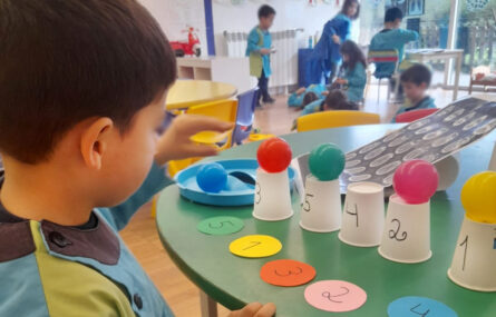 Uma criança brinca com pequenas bolas coloridas que coloca em cima de copos invertidos. Na mesa estão também pequenos círculos de papel numerados. Ao fundo outras crianças brincam. Os rostos das crianças foram digitalmente desfocados.
