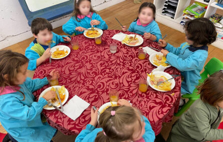 Sete crianças sentadas a comer (numa mesa redonda). Em cima da mesa estão pratos com comida. Os rostos das crianças foram digitalmente desfocados.